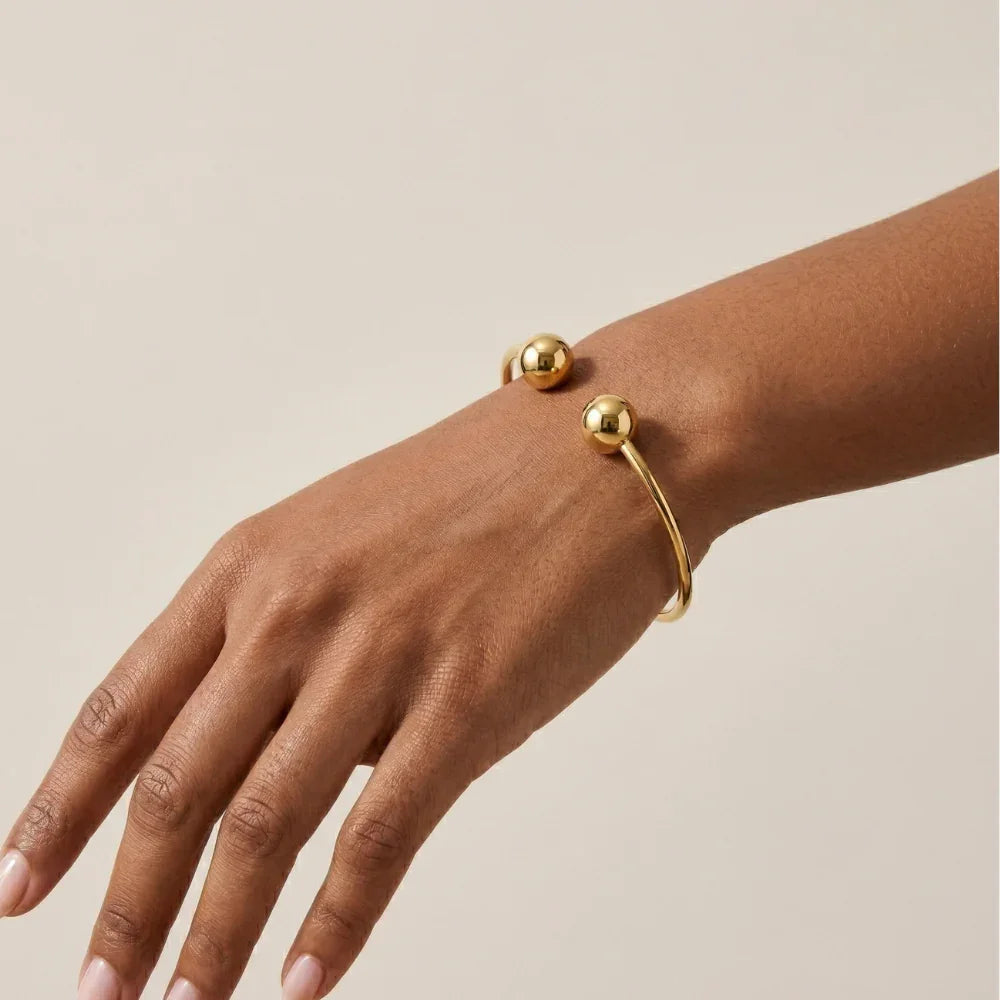 Bracelete Bola Rígido dourado, aberto, com duas esferas lisas nas pontas, usado no pulso sobre fundo claro