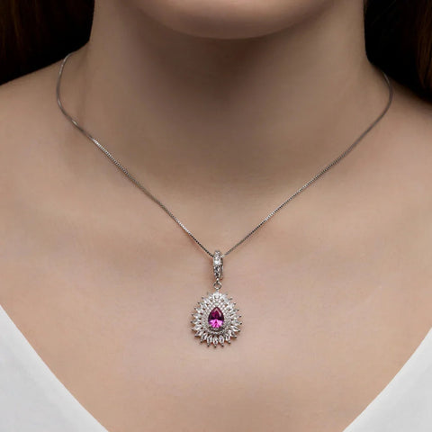 Pingente Zircônia Central Cravejado com pedra rosa em gota e contorno de zircônias brancas, em corrente fina prateada sobre o colo, com blusa branca de decote V
