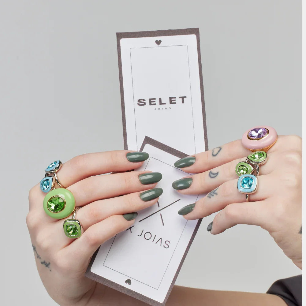 Anéis coloridos com pedras brilhantes da Selet Joias, exibidos em mãos com unhas verdes e cartão da marca
