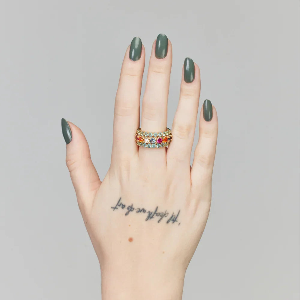 Anel colorido com pedras preciosas em mão com unhas escuras e tatuagem